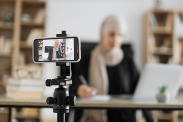 Focus on screen of phone of cheerful young muslim woman in hijab recording creative video blog for her social networks, media for followers while working with laptop and writing report.