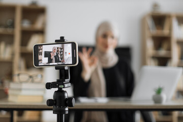 Naklejka premium Focus on screen of phone of happy arab woman sitting at desk with books, notes and laptop talking with followers. Female tutor working at home during distance learning and showing sign ok.