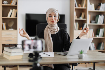 Muslim woman freelancer sitting at table with closed eyes recording blog how to relieving stress by meditation at workplace. Concept of relaxation and harmony, no stress free relief at work.