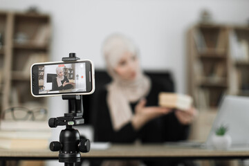 Focus on phone of muslim woman in casual wear and hijab recording video on smartphone while unpacking gift boxes. Female blogger sharing her emotions with her subscribers in social networks.