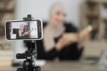 Focus on phone of happy muslim female blogger unpacking gift boxes presents from companies and doing live streaming. Arab woman using smart phone camera for creating content.