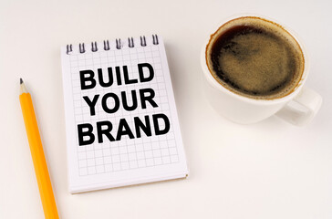 On a white surface, a cup of coffee, a pencil and a notepad with the inscription - Build Your Brand
