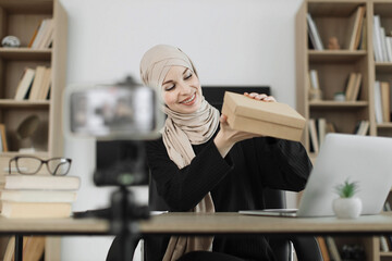 Pretty muslim woman in casual wear and hijab recording video on camera while unpacking gift boxes. Female blogger sharing her emotions with her subscribers in social networks.