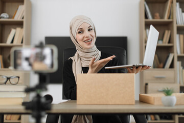 Pleasant arab woman filming video on modern phone camera while opening parcel box with new wireless laptop. Concept of people, technology and blogging.