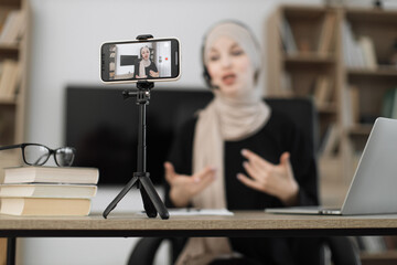 Charming muslim woman in hijab and headset talking and gesturing while recording video on modern...