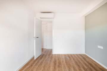 A wide empty bright room with one soft gray wall. The floor is light wood parquet. Above the open door is an air conditioner to regulate the temperature inside the room.