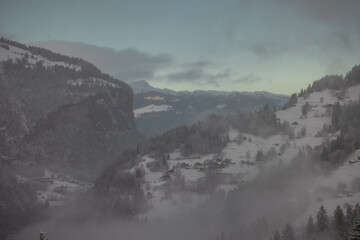 Looking down towards the Lauterbrunnen valley from Wengen area with low clouds and some haze in the air during winter time. Some cottages visible..