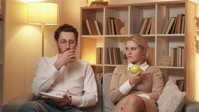 Unhealthy food. Addicted woman. Diet problem. Happy man enjoying eating sweet doughnut while envious wife taking over and changing to apple in light shadow room interior.