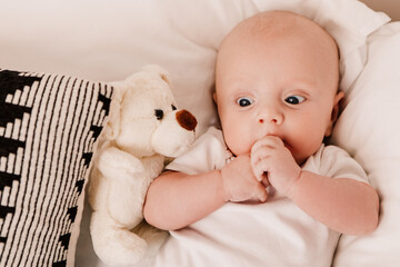 Little boy funny cute child baby lying on bed pillows with fluffy toy. Playful toddler with bulging eyes having fun, making faces grimaces. Happy childhood, family concept
