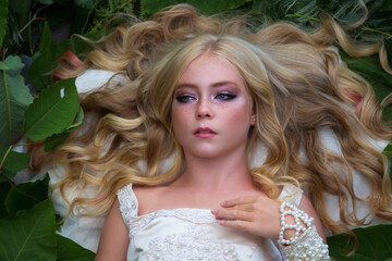 Fototapeta premium Close-up portrait of beautiful preteen girl with curly blond hair and facial frecles wearing white dress and lying on her back on green leaves. Serene people. Selective focus. Beauty and Fashion theme