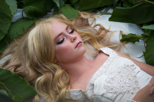 Close-up portrait of beautiful preteen girl with blond hair and facial frecles lying on her back on green leaves with closed eyes. Serene people. Selective focus. Beauty and Fashion theme