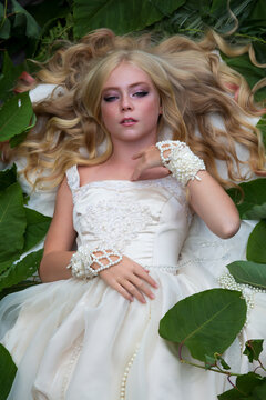 Close-up portrait of beautiful preteen girl with curly blond hair and facial frecles wearing white dress and lying on her back on green leaves. Serene people. Selective focus. Beauty and Fashion theme