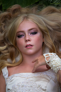 Close-up portrait of beautiful preteen girl with curly blond hair and facial frecles wearing white dress and lying on her back. Serene people. Selective focus. Beauty and Fashion theme.