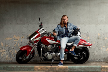Plakat Lifestyle of a motorcyclist, a handsome biker with long hair on a red classic bike in an urban landscape.