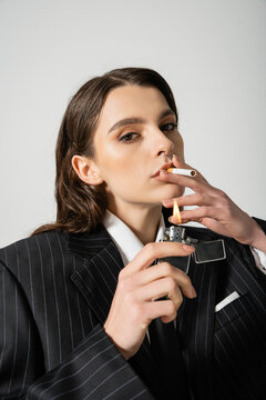 fashionable woman with makeup and piercing lighting cigarette and looking at camera isolated on grey.