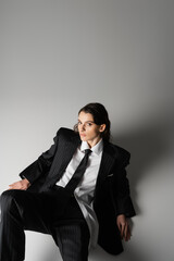 high angle view of brunette woman in black oversize suit and white shirt sitting and looking at camera on grey background.