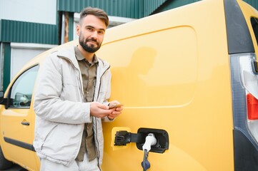 Fototapeta na wymiar bearded caucasian man standing near an electric car that is charging and making time adjustments on a smartphone.