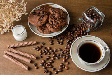 A cup of coffee with chocolate cookie on a dark wooden table. Spilled coffee beans. Milk in a glass jar. Top view.
