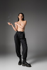 full length of shirtless and sexy woman in black pants and rough boots posing with cigarette and looking at camera on grey.