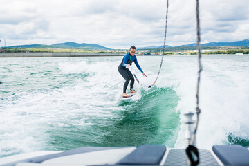 Watersport concept. Young athletic woman learning wakesurfing and perfecting tricks. Female in wetsuit riding the waves using of tow rope behind a boat on sunny day.