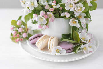 Obraz na płótnie Canvas Beautiful composition with delicious French macarons and spring flowers in a white cup. Sweet dessert, early spring white and pink flowers, wedding decor, bride morning. Greeting card Mother's Day