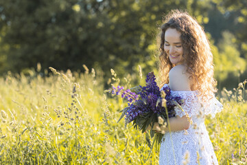 Portrait of beautiful happy smiling young girl in a white dress with curly hair, straw hat, with picnic and bouquet of purple wild flowers on a meadow. Summertime, golden hour, sunset, wellbeing