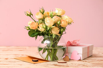 Glass vase with beautiful rose flowers, envelope and gift box on light wooden table near pink wall