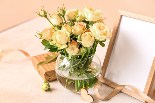 Vase with beautiful rose flowers, gift box and empty picture frame on table near color wall