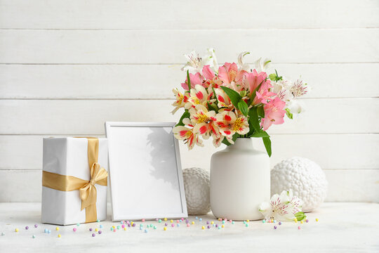 Vase with beautiful alstroemeria flowers, empty picture frame, gift and decor on table near light wooden wall. Mother's day celebration