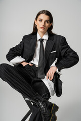 stylish brunette woman in elegant oversize suit sitting with hand in pocket while looking away isolated on grey.