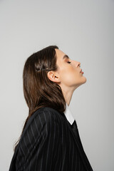 side view of young brunette woman in black and striped blazer isolated on grey.