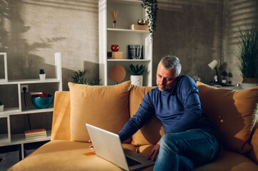 Middle aged man using smartphone and a laptop while sitting on a sofa at home