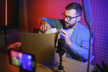 Handsome young man gesturing while unpacking parcel, excited, clenches fists in triumph. Joyful male vlogger. Concept of people and social networks.