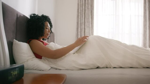 Cinematic shot of young woman of color reading book while relaxing with pleasure in comfortable bed on sunny morning in bedroom at home. Concept of leisure, education, rest, comfort, relaxation 4K