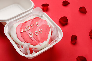 Plastic lunch box with heart-shaped bento cake and rose petals on red background. Valentine's Day celebration