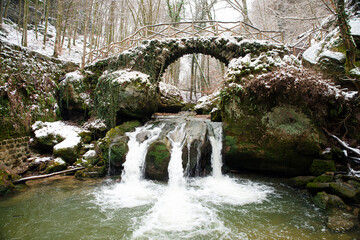 Scheissendempel waterfall, river Black Ernz with stone bridge covered with snow, Mullerthal trail in Waldbillig, Luxembourg in winter
