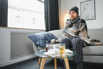 Fototapeta na wymiar Sick bearded man who has bad cold or seasonal flu sitting on couch at home. Guy with fever wearing warm plaid shivering with worried face expression.