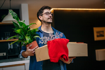 University student standing, holding a plant and cardboard box, unpacking and arranging things in a...