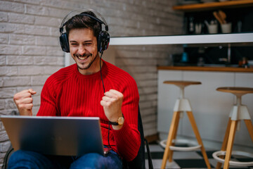 Handsome gamer celebrating online tournament victory from home. Excited man making yes gesture enjoying triumph. Overjoyed gamer winning in shooter battle in the headset.