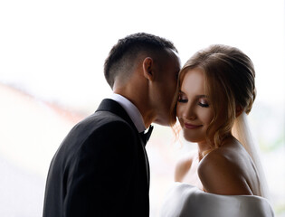 Close view of pretty woman with stylish hairdo, dressed in white gown with naked shoulders, smiling and looking away while faceless groom kissing her face during wed celebration