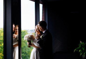Side view of incognito bride groom in trendy black suit with modern watch on hand, kissing girl face and embracing her shoulder while she standing near large window in long dress, holding bouquet