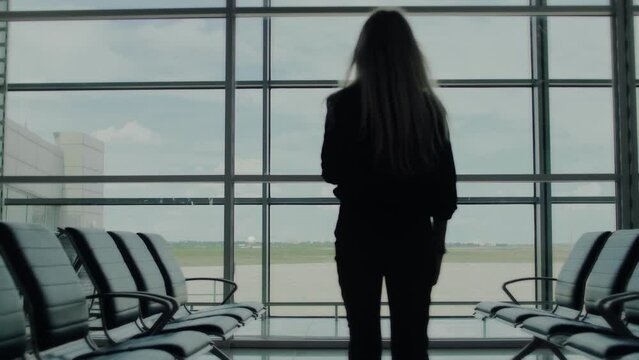 A blonde woman in a blue shirt strolls through an empty airport lounge, with chairs and airport field in the background