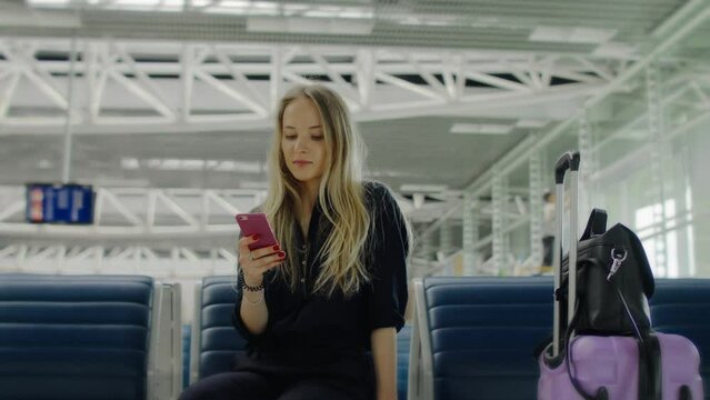 Cheerful young blonde woman sitting in airport hall with suitcase and phone. 4K footage