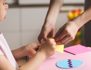 The hands of a little caucasian girl hold a yellow sticker, and mom's hands help peel off a strip for felt eggs.