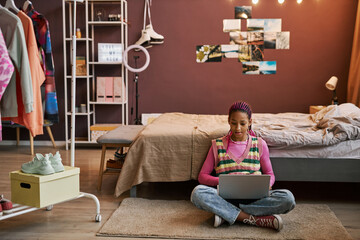 Black teenage girl sitting on floor in pink room interior using laptop