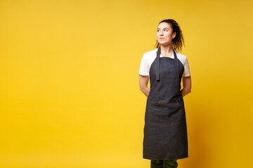 girl waiter in uniform stands with her hands behind her back and looks away, barista woman in denim apron - obrazy, fototapety, plakaty