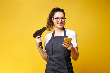 young girl hairdresser in uniform holds hair dryer and uses smartphone on yellow background