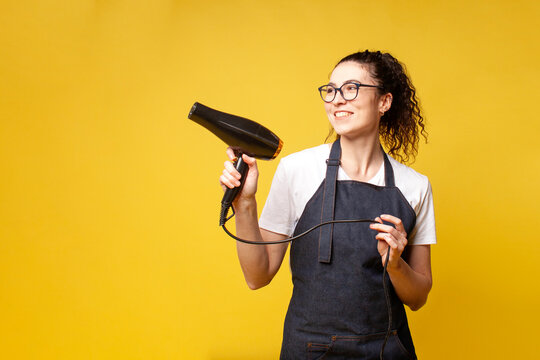 young girl hairdresser in uniform holds a hair dryer and smiles on a yellow background, woman stylist in an apron