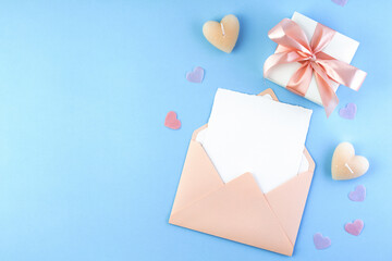 Pink envelope with blank white paper card inside, Valentines gift, hearts on pastel blue background. Happy Valentine's Day flat lay composition.