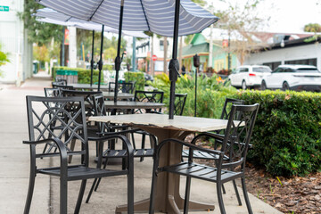 Multiple empty black plastic tables with square glass tops and black chairs at a sidewalk cafe patio of a restaurant. The outdoor restaurant has large white sunshade umbrellas. 